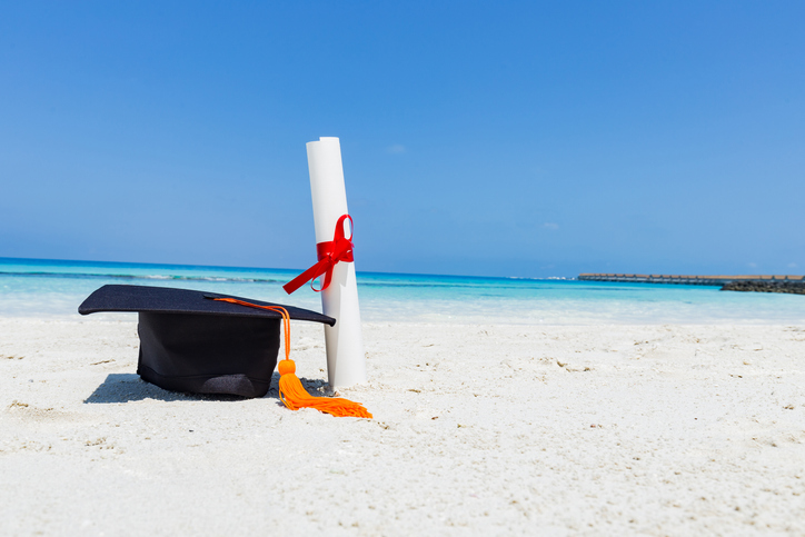 A photo of a graduation cap and diploma on a beach.