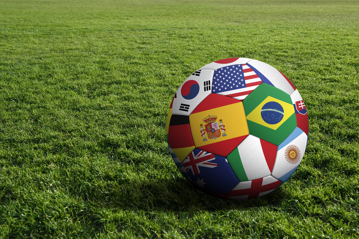 A picture of a soccer ball with international flags painted on it sitting on a green field.